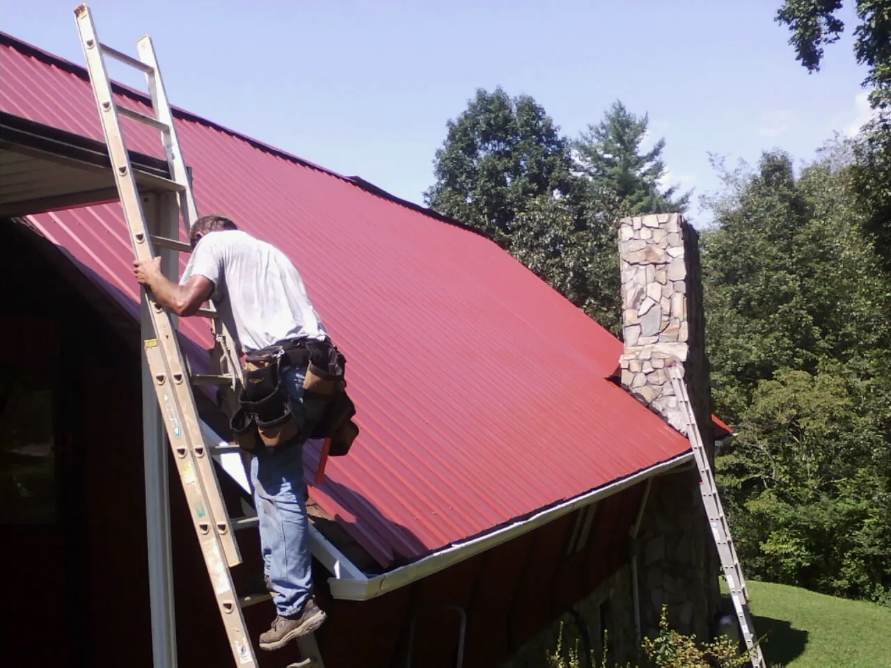 Roofer on a ladder beside a red metal roof and stone chimney — Soffit & Fascia Repair by Roofing Brothers in Poultney